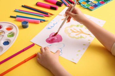 Sarı arka planda fırça, boya ve kalemlerle kalbi çizen bir çocuk.