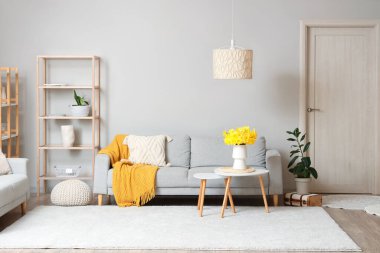 Rahat bir kanepe, raf ünitesi ve oturma odasında vazo dolusu narsist çiçekli kahve masası.