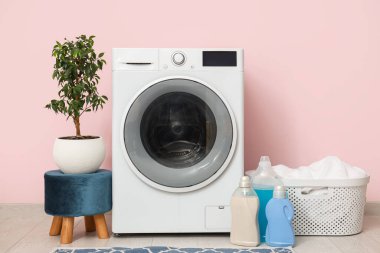 Pembe duvarın yanında sepetli, çiçekli ve deterjanlı modern çamaşır makinesi.