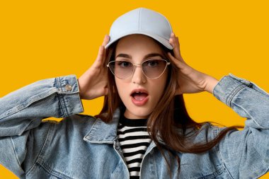 Şok olmuş, sarı arka planda güneş gözlüklü ve şapkalı genç bir kadın.