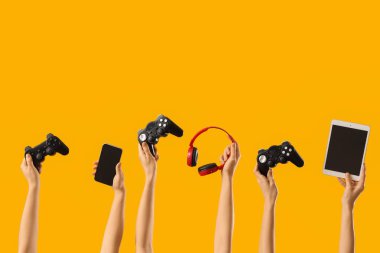 Sarı arka planda oyun pedleri, cep telefonu, tablet bilgisayar ve kulaklıkları olan kadın eller