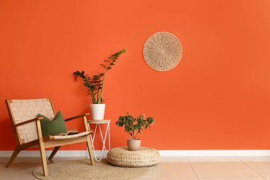 Hasır pouf ve turuncu duvarın yanındaki bitkilerle dolu rahat bir koltuk.