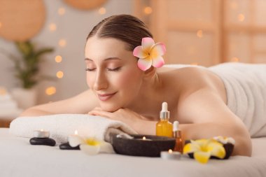 Pembe tesisat çiçekleri, kozmetik yağı ve mumlarla kaplıca salonundaki kanepede uzanan güzel, genç ve mutlu bir kadın.