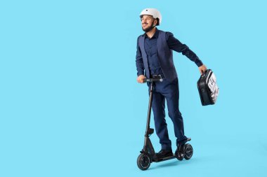 Mavi arka planda elektrikli scooter kullanan takım elbiseli ve kasklı genç adam.