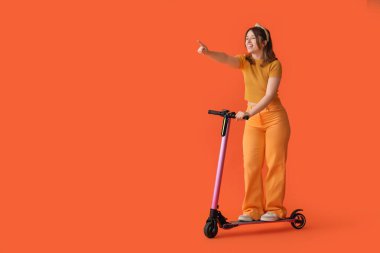 Elektrikli scooter kullanan mutlu genç bir kadın ve turuncu arka plandaki bir şeyi işaret ediyor.