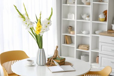 Mutfaktaki yemek masasının üzerinde gladiolus çiçekleri, kitaplar ve fincanlar olan vazo.