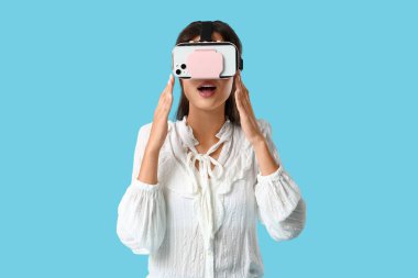 Modern VR gözlüklü, mavi arka planda cep telefonu olan şok olmuş genç bir kadın.