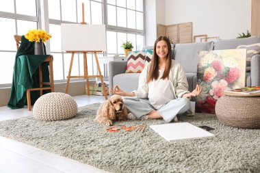 Hamile sanatçı köpekle yerde oturuyor ve odada meditasyon yapıyor.