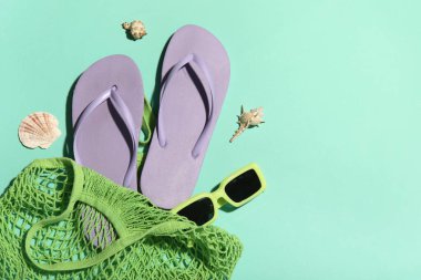 Parmak arası terlik, deniz kabuğu ve turkuaz arka planda güneş gözlüğü olan bir çanta.