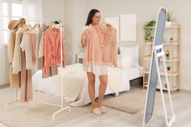 Genç bir kadın yatak odasında aynanın yanında şık bir bluz deniyor.