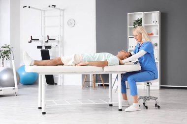 Rehabilitasyon merkezindeki kanepede yatan genç adamla çalışan kadın fizyoterapist.