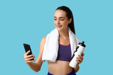Güzel, genç, sportif, mutlu bir kadın. Mavi arka planda bir şişe su, havlu ve cep telefonu var.