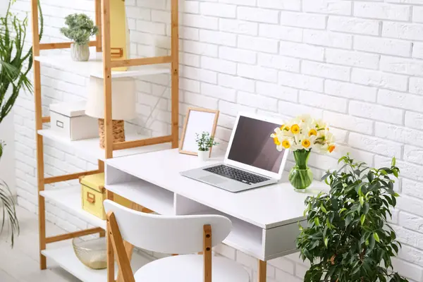 Ofisteki beyaz tuğla duvarın yanında bir buket narsisli çiçek ve dizüstü bilgisayarla raf ve işyeri.