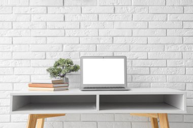 Ofiste beyaz tuğla duvarın yanında modern dizüstü bilgisayarı ve ev bitkisi olan şık bir iş yeri.