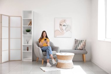 Odadaki koltukta otururken cep telefonu kullanan genç bir kadın.