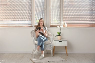 Orkide çiçeğinin yanındaki koltukta otururken cep telefonu kullanan genç bir kadın.