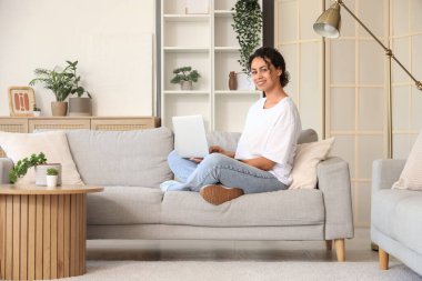 Genç Afro-Amerikalı kadın evdeki gri koltukta dizüstü bilgisayar kullanıyor.