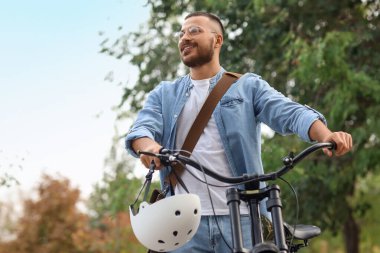 Kasklı ve bisikletli genç adam sonbahar günü parkta yürüyor.