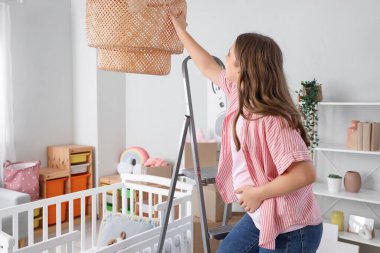 Genç hamile kadın çocuk odasında tamir sırasında lamba asıyor.