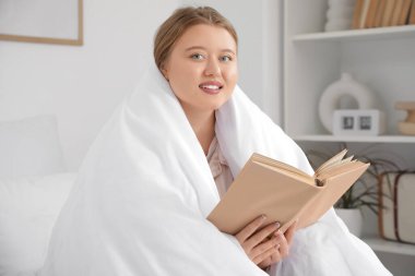 Güzel, genç, mutlu bir kadın battaniye içinde evde kitap okuyor.