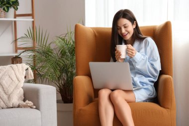 Kahve ve dizüstü bilgisayarlı mavi pijamalı genç bir kadın evde koltukta çalışıyor.