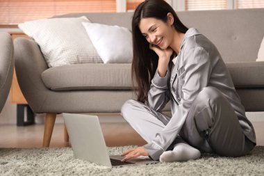 Gri pijamalı genç bir kadın evde dizüstü bilgisayarla çalışıyor.