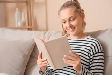 Evdeki kanepede kitap okuyan güzel sarışın kadın.