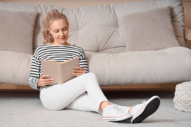 Evdeki halıda kitap okuyan güzel sarışın kadın.