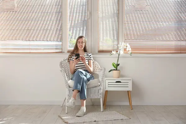 Orkide çiçeğinin yanındaki koltukta otururken cep telefonu kullanan genç bir kadın.