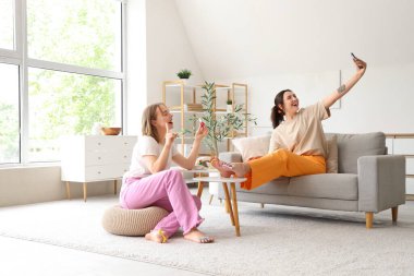 Genç kadınlar pedikür yapıyor ve oturma odasında selfie çekiyor.