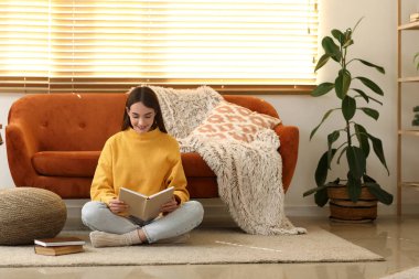 Genç bir kadın oturma odasında oturmuş kitap okuyor.