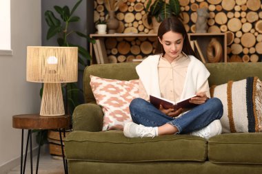 Oturma odasındaki kanepede oturmuş kitap okuyan güzel genç bir kadın.