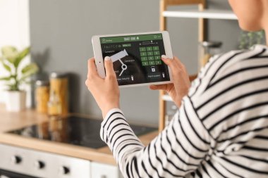 Mutfakta akıllı ev güvenlik sistemi kontrol paneli kullanan genç bir kadın.