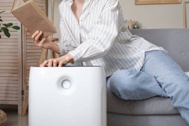 Genç kadın kanepede oturuyor ve oturma odasındaki modern nemlendiriciyi açıyor.