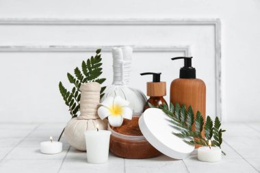 Farklı kozmetik ürünleri, bitkisel poşetler, eğrelti otu dalları, erik çiçeği ve beyaz fayans masasında yanan mumlar içeren spa malzemeleri.