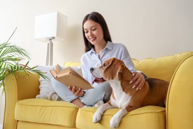 Oturma odasındaki sarı kanepede sevimli Beagle köpeğiyle kitap okuyan genç bir kadın.