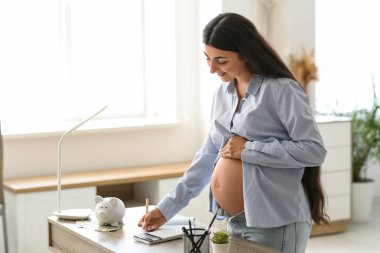 Evdeki masanın üzerine not defterine yazan hamile bir kadın. Annelik Faydası konsepti