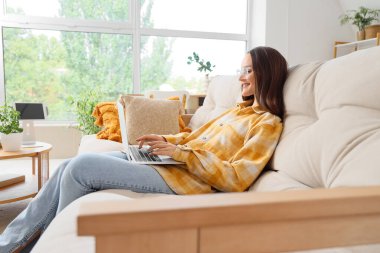 Evdeki kanepede dizüstü bilgisayar kullanan gülümseyen genç kadın.