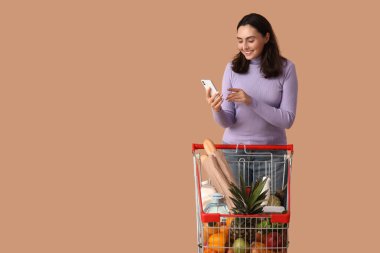 Güzel, genç, mutlu bir kadın, alışveriş arabası ve bej arka planda cep telefonu var.