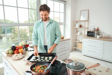 Yakışıklı sakallı adam mutfaktaki yemek tepsisine kabak dilimleri koyuyor.