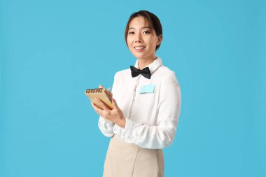 Mavi arka planda not defteri olan gülümseyen genç Asyalı garson.