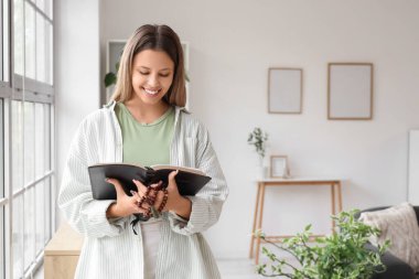 Dua boncukları olan genç bir kadın. Evinin penceresinin yanında İncil okuyor.