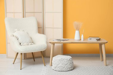 Sari duvarin yakinlarinda, katlanabilir ekran, koltuk, puf ve masa olan bir oda.