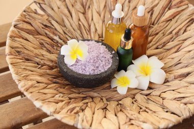 Üzerinde kozmetik ürünler şişeleri, deniz tuzu ve ahşap masa üzerinde tropikal çiçekler olan hasır bir tabak, yakın plan.