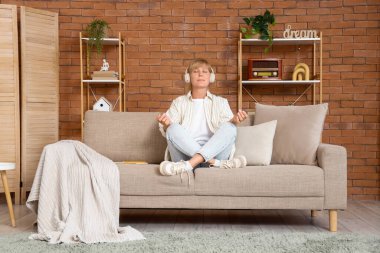 Kulaklıklı olgun bir kadın evdeki kanepede meditasyon yapıyor.