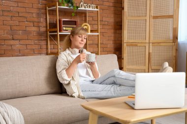 Evde kanepede oturan çay ve dizüstü bilgisayarı olan olgun bir kadın.