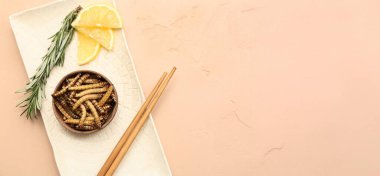 İçinde kızarmış kurtçuklar, yemek çubukları, biberiye ve bej arka planda limon dilimleri olan bir kase.