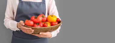 Gri arka planda farklı taze domateslerle dolu hasırdan kaseyi tutan bir kadın. Mesaj için yeri var, yakın plan.