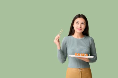 Düşünceli genç Asyalı kadın elinde yemek çubukları ve lezzetli suşili tabakla yeşil arka planda rulo yapıyor.