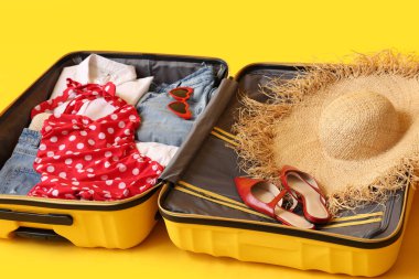 Sarı arka planda kadın kıyafetleri, şapka ve ayakkabıları olan bavulu aç. Seyahat kavramı